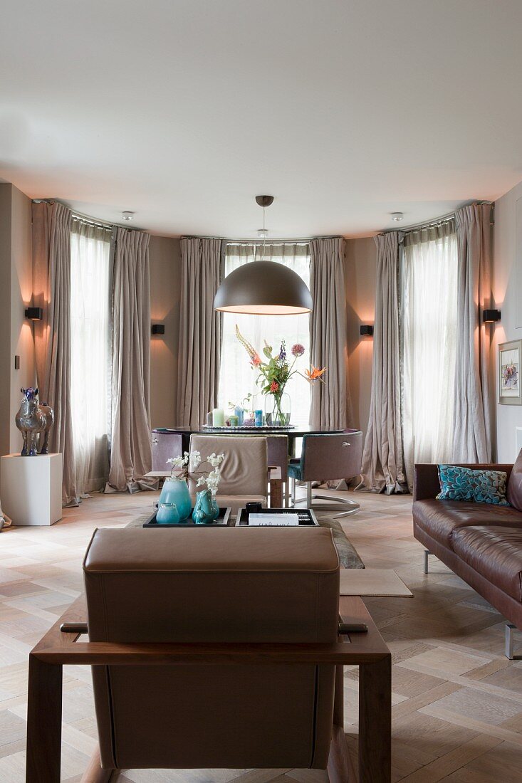 Eleganter Loungebereich mit Ledersitzmöbeln in Braun, im Hintergrund halbrunder Erker mit Wandbeleuchtung zwischen Fenster mit bodenlangem Vorhang