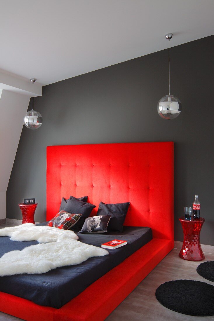 Dramatisch gestaltetes Schlafzimmer, Tierfell und Matratze auf Bodenpolster mit hohem Kopfteil in Rot an dunkelgrauer Wand