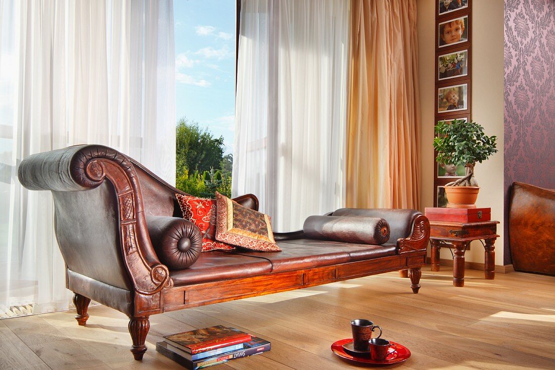 Elegante, antike Chaiselongue vor Panoramafenster mit luftigem Vorhang in traditionellem Ambiente