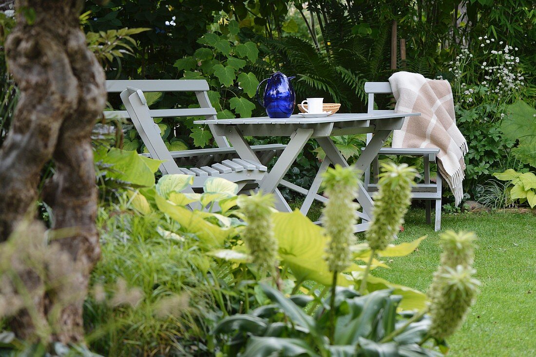 Tisch und Stühle aus grau lackiertem Holz auf Rasen im Garten, umgeben von Grünpflanzen