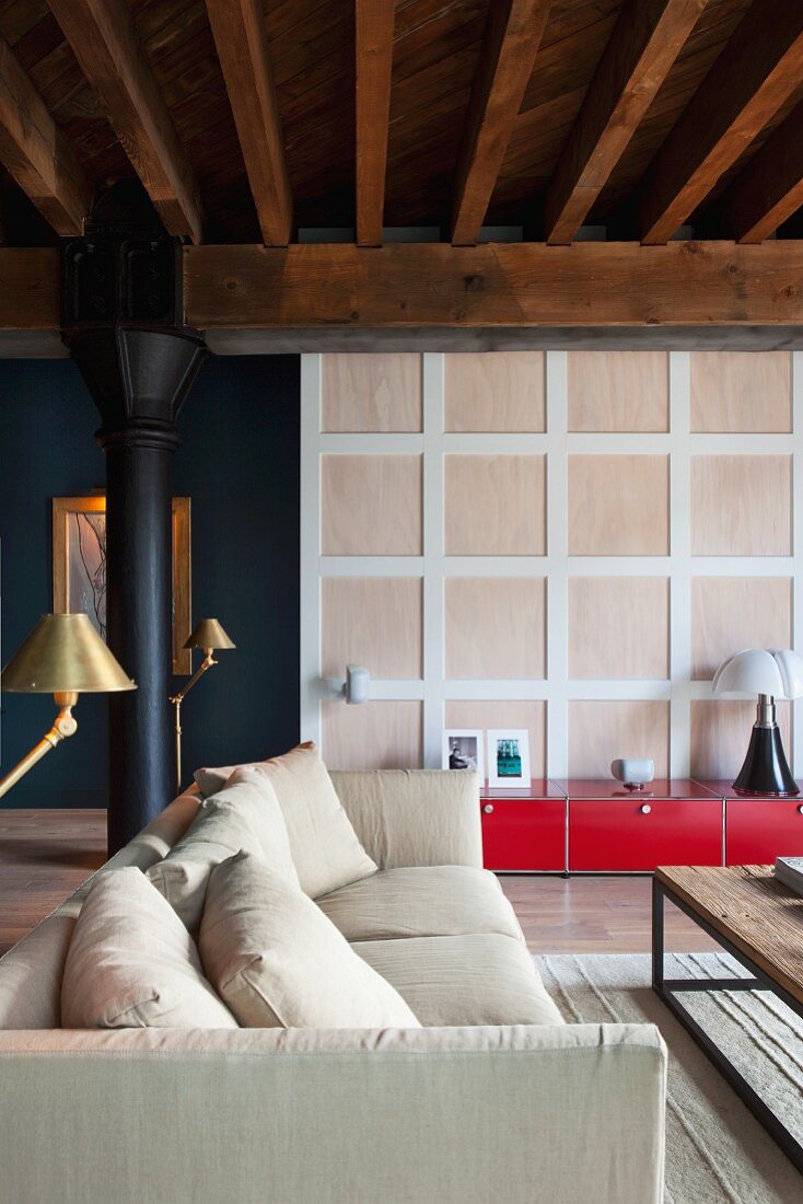 Sitzbereich mit Sofa und Messingleuchten in Loft mit gusseisernen Stützen und Holzbalkendecke; rotes Lowboard vor Raumteiler Kassettenwand