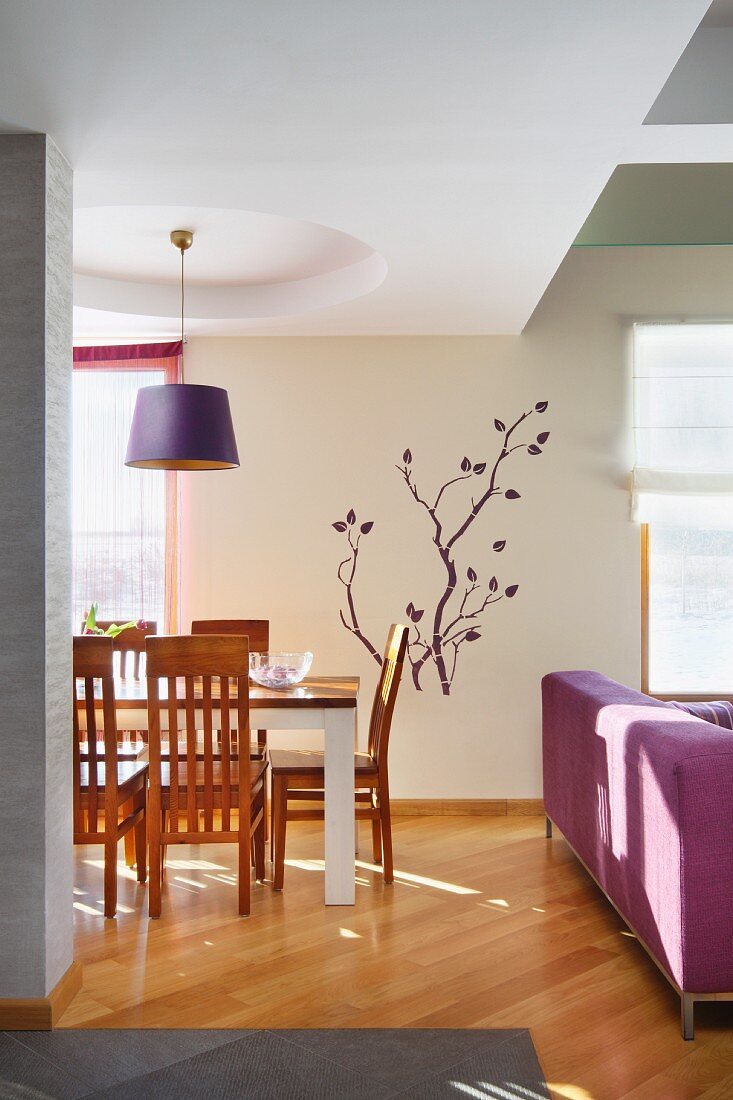Eleganter Essplatz mit Holzstühlen unter Hängeleuchte mit lila Schirm, dahinter Schablonenmalerei mit Zweigenmotiv an Wand, seitlich teilweise sichtbare Couch