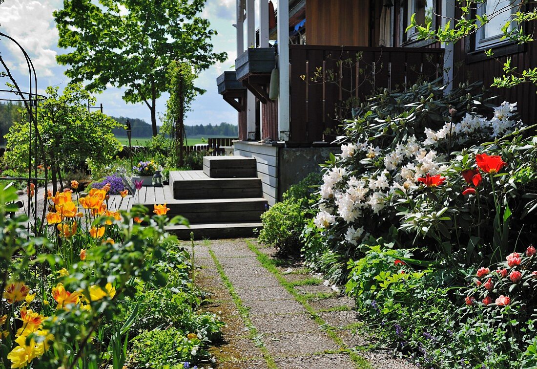 Gepflasterter Weg zwischen Blumenbeeten in sommerlichem Garten, im Hintergrund Treppe vor Veranda eines Holzhauses