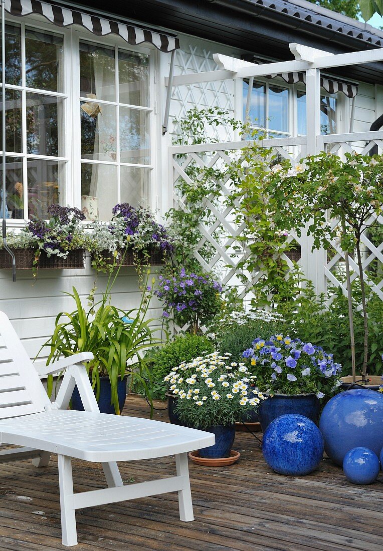 Weisser Liegestuhl neben Blumen in blauen Übertöpfen auf Holzterrasse vor weißem Holzhaus