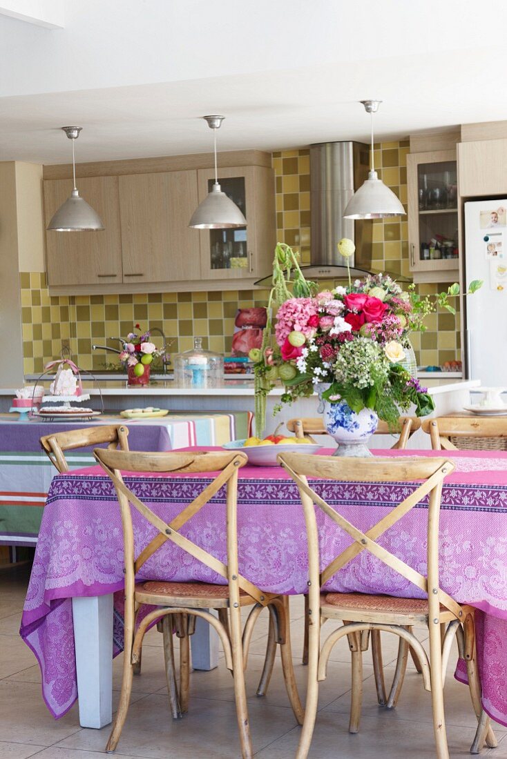 Blumenstrauss auf Tisch mit bunter Tischdecke und alte Holzstühle in moderner Küche, Hängeleuchten in Reihe über Theke