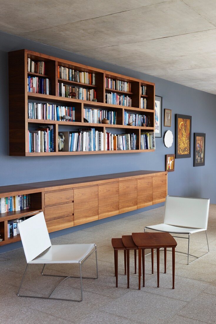 Moderne Stühle mit hellem Bezug und mehrteiliges Beistelltisch-Set aus Holz vor Sideboard unter aufgehängtem Bücherregal an grau getönter Wand