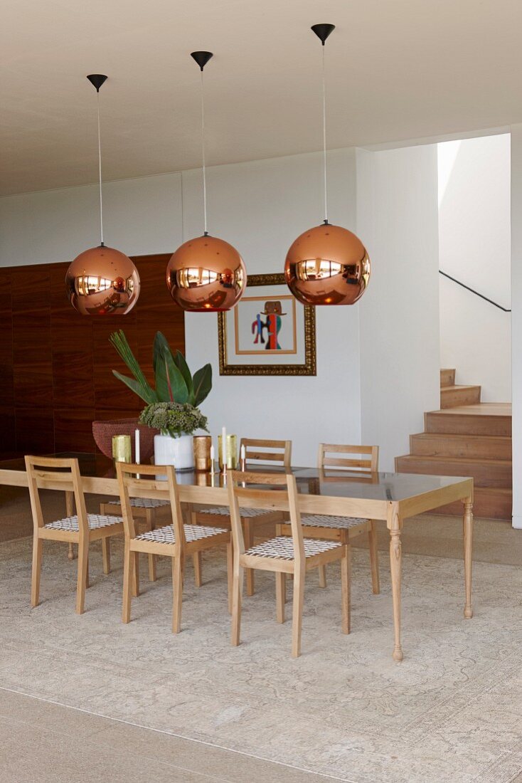 Lange Designertafel und Stühle aus hellem Holz, unter Pendelleuchten mit kupferfarbenem Schirm, in modernem Essbereich