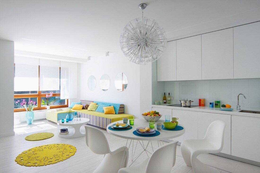 Offener Wohnraum in Weiß, Essplatz mit Klassiker Schalenstühlen gegenüber weisser Einbauküche, gelbe, runde Teppiche als Farbtupfer