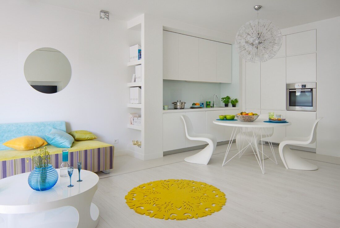 Lounge- und Essbereich mit Einbauküche in offenem, weißem Wohnraum, gelber, runder Teppich als Farbakzent auf weißem Boden