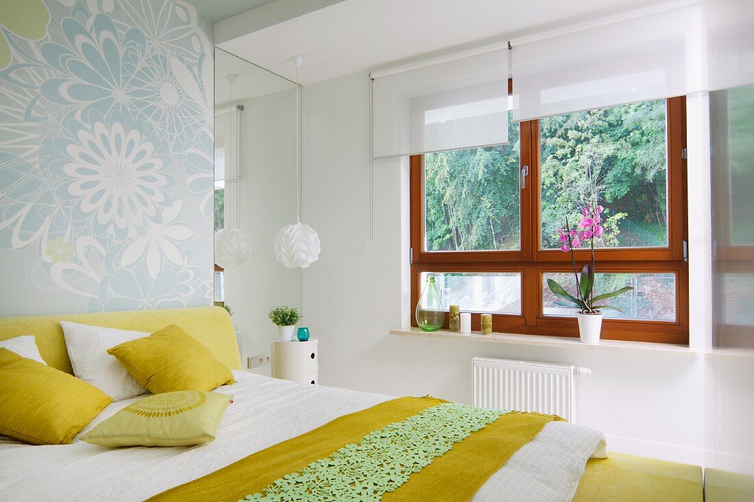 Schlafzimmer mit Doppelbett, Bettwäsche und Kissen in Weiß und Gelb, gegenüber Holzfenster mit weissen Rollos und Gartenblick
