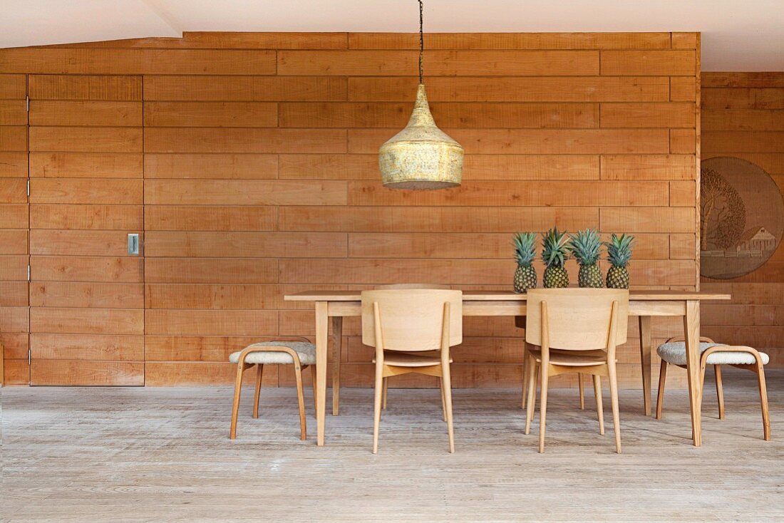 Stühle, Hocker und Tisch aus hellem Holz, darauf frische Ananas, oberhalb Vintage Pendelleuchte, holzverkleidete Wand