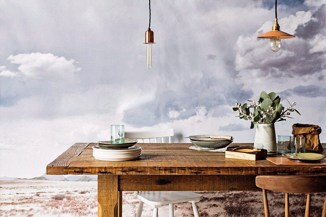 Rustikaler Tisch mit Tellerstapel und Vase, unter Vintage Pendelleuchten, im Hintergrund Bild mit grauer Wolkenstimmung