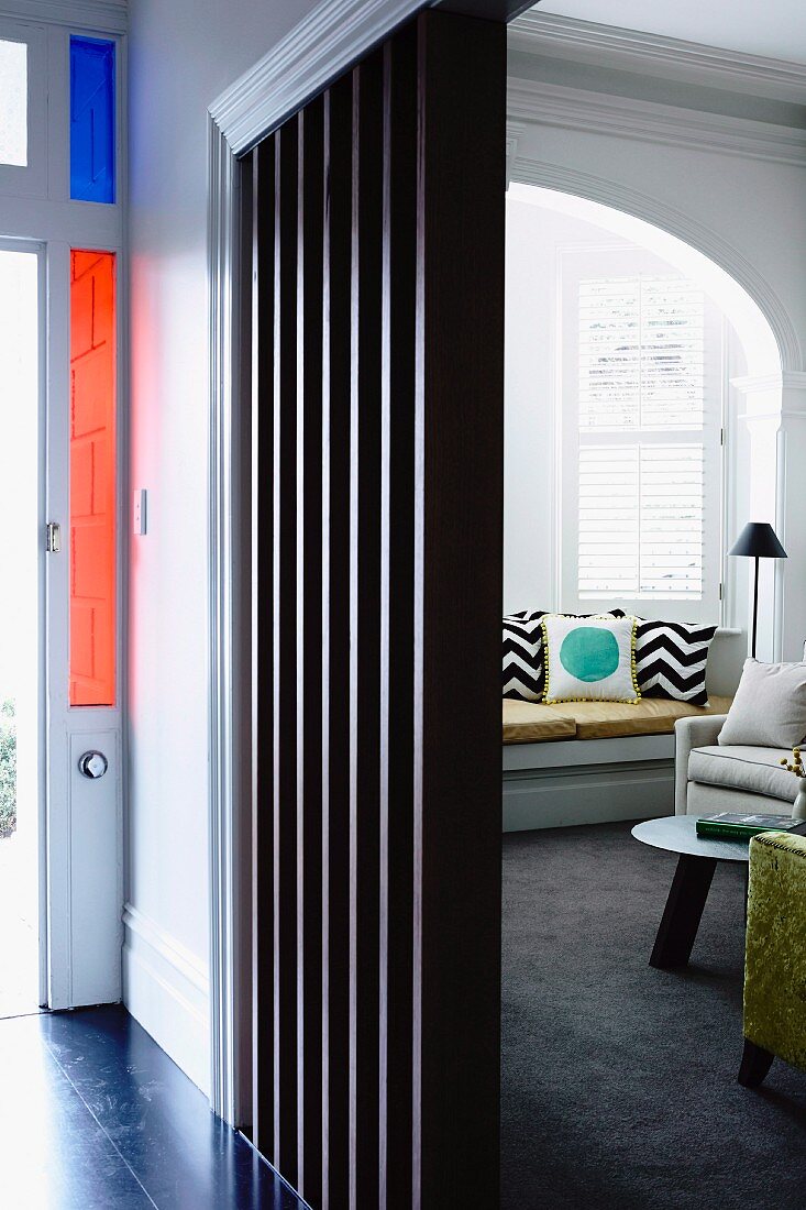 Blick auf farbige Fensterelemente der Haustür; Wohnzimmer mit Sofa im Fenstererker, vorne schwarze Holzlamellen als Raumteiler