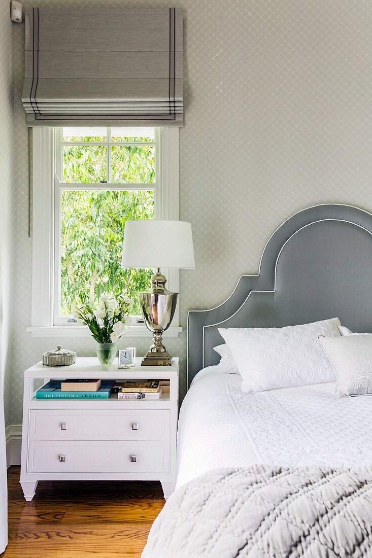 White bedside cabinet below window with Roman blind next to bed with curved headboard upholstered in grey