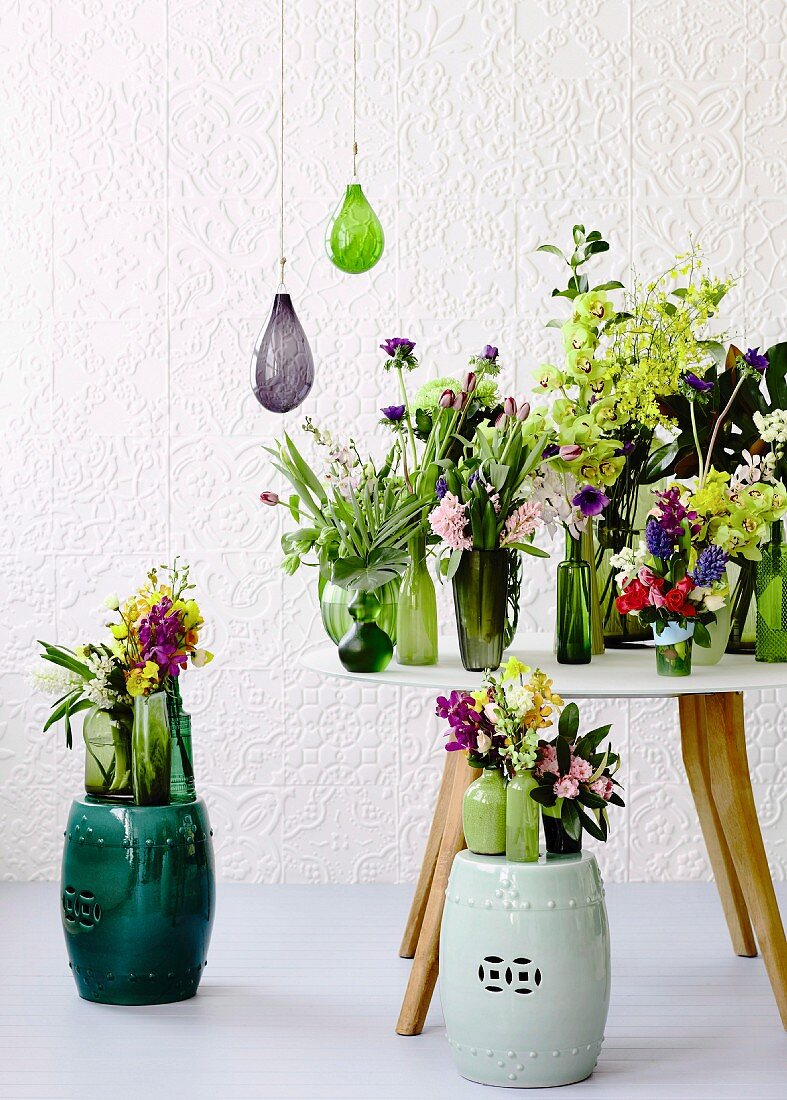 Spring bouquets in green glass vases on table and on ceramic stools against white structured wallpaper