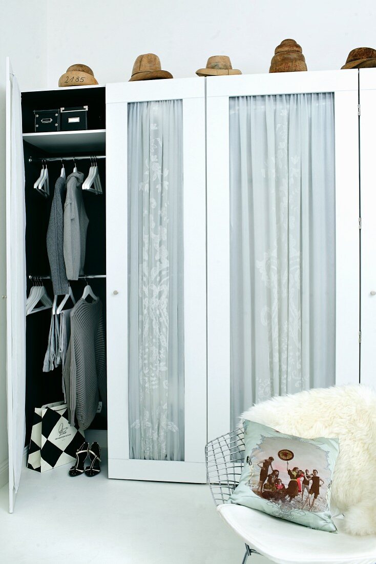Kleiderschrank mit Glastüren und Vorhang, oberhalb Hutformen, im Vordergrund Klassikerstuhl mit Tierfell und Kissen