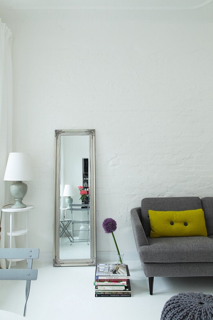 Standspiegel an geweisselter Ziegelwand zwischen Beistelltisch mit Leuchte und grauem Sofa