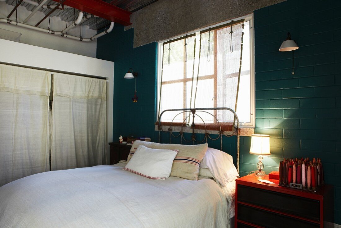Bett mit Vintage Metallgestell vor Fenster, seitlich Tischleuchte auf rotem Nachtkästchen vor grün getünchter Ziegelwand