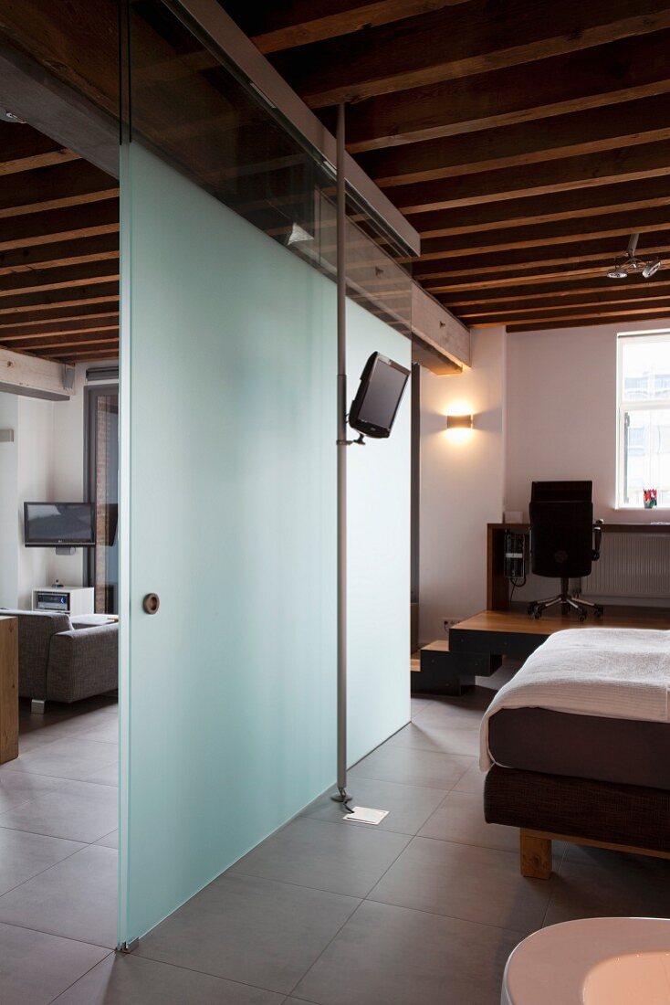 Glas-Raumteiler mit Schiebetür zwischen Schlafbereich und Wohnen in eleganter Loftwohnung mit rustikaler Holzbalkendecke
