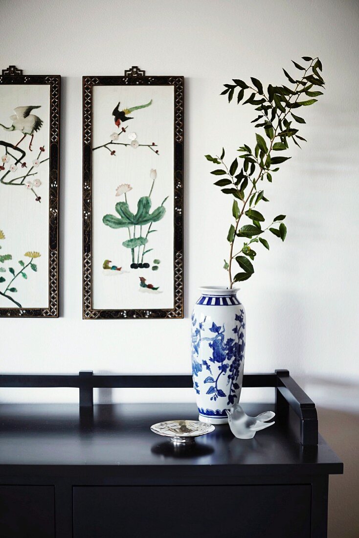 White and blue painted porcelain vase on black sideboard below framed pictures with Oriental motifs