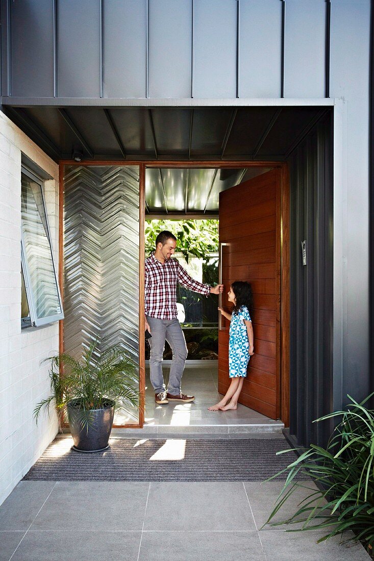 Vater und Tochter im überdachten Eingangsbereich eines Wohnhauses mit grauschwarzer Metallfassade; neben der offenen Tür ein verglastes Element mit Zickzack-Struktur