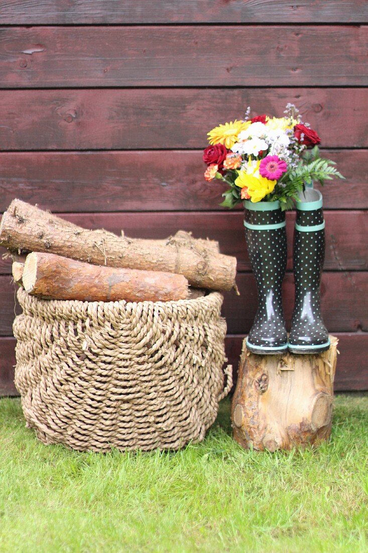 Korb mit Holz neben Gummistiefeln und Blumen
