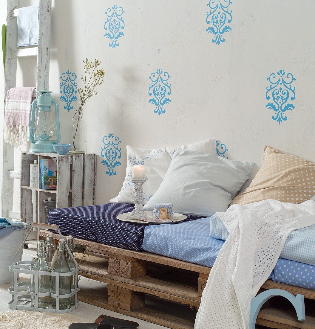 Selbstgebautes Tagesbett aus gestapelten Holzpaletten, Polster und Kissen, vor Wand mit hellblauer Ornament-Schablonenmalerei
