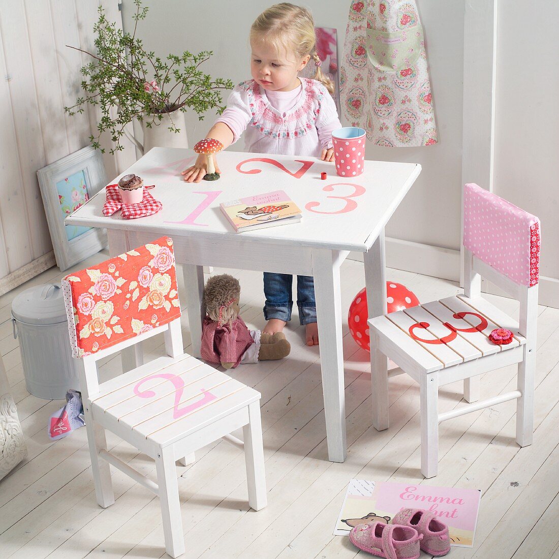 Little girl sitting at white, children's table painted with numbers; chairs with patterned, fabric backrest covers