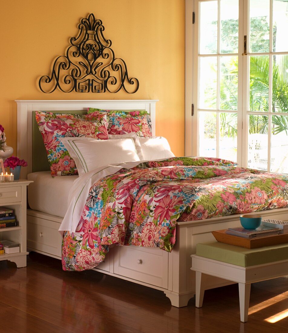 Bedroom with yellow walls, white bed and floral bed linen next to French doors overlooking garden