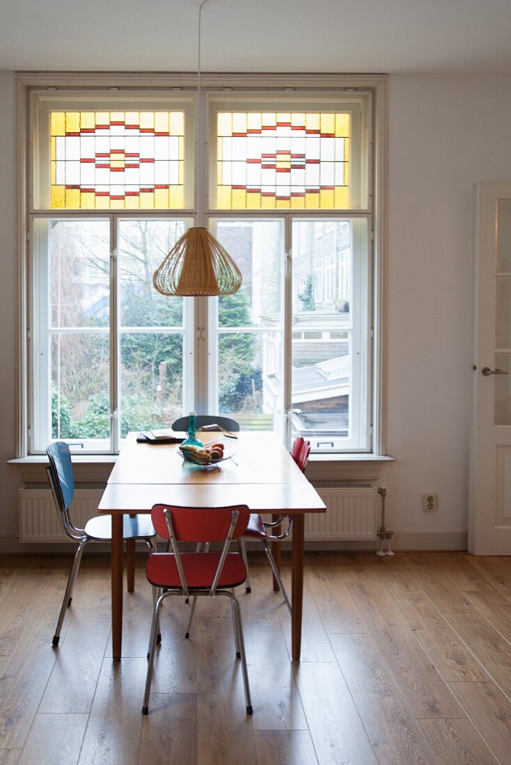 Retro chairs around kitchen table in front of window with stained glass upper panes in simple dining room