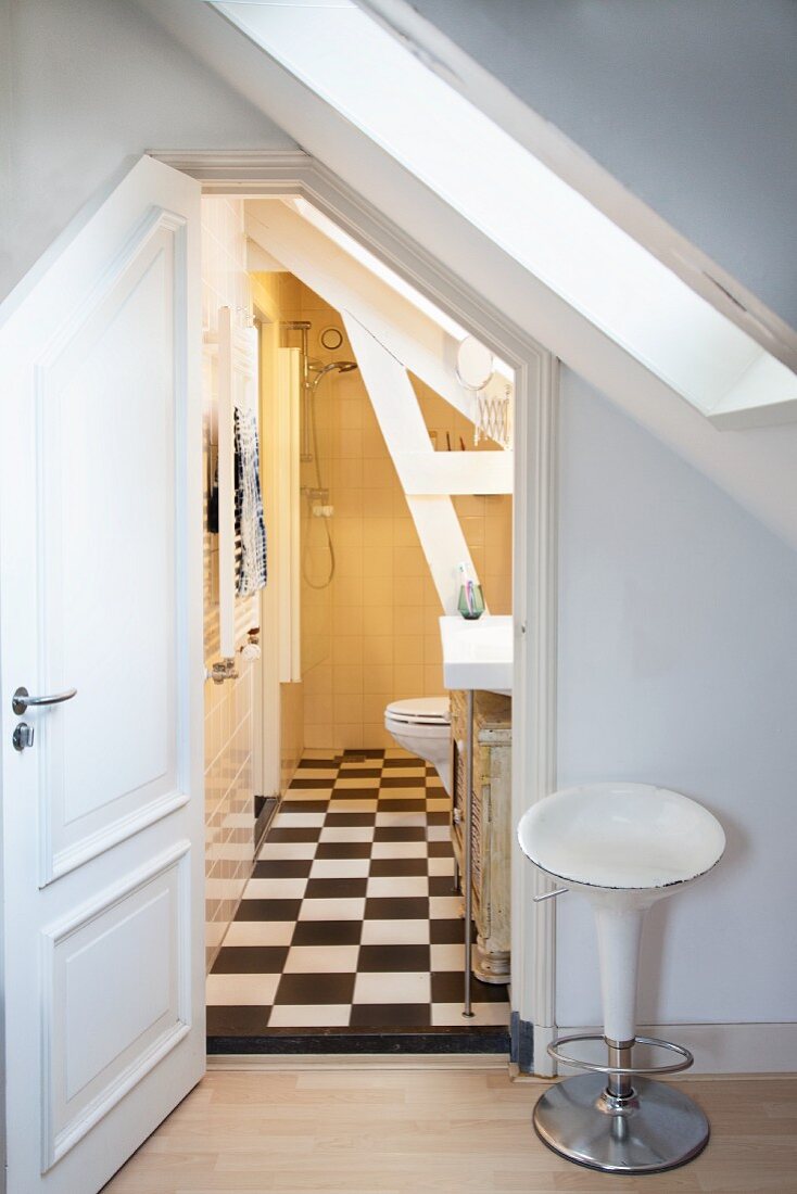 Vintage Barhocker neben offener Tür und Blick in ländliches Bad unter dem Dach, schwarz-weisser Schachbrettmusterboden