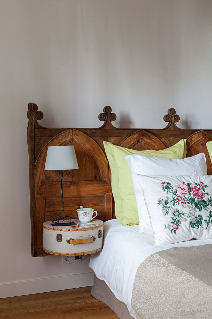 Bed with antique, carved wooden headboard; table lamp on small, retro case mounted on headboard as bedside table