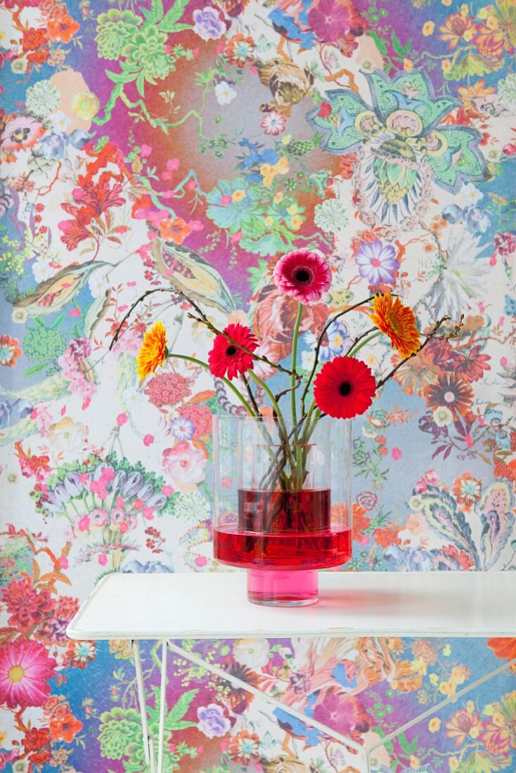 Gerbera in Glasvase mit roter Flüssigkeit vor floralem Hintergrund