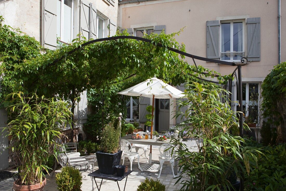 Sonnenbeschienener Terrassenplatz unter idyllisch berankter Metallpergola im Innenhof eines französischen Landhauses