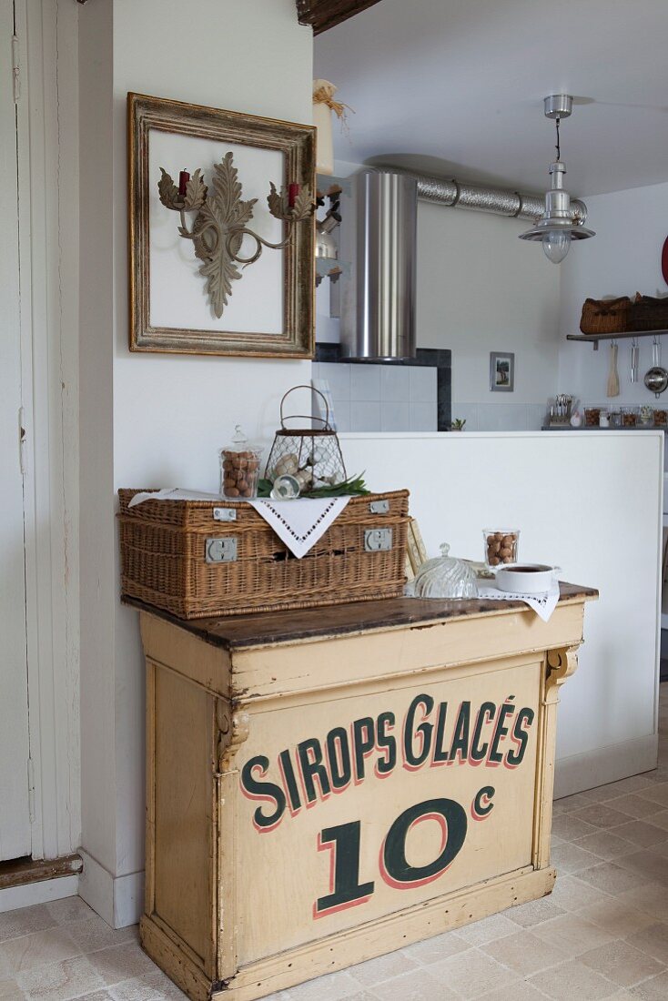 Vintage cabinet and wicker hamper next to white, half-height wall; stainless steel extractor hood in kitchen in background