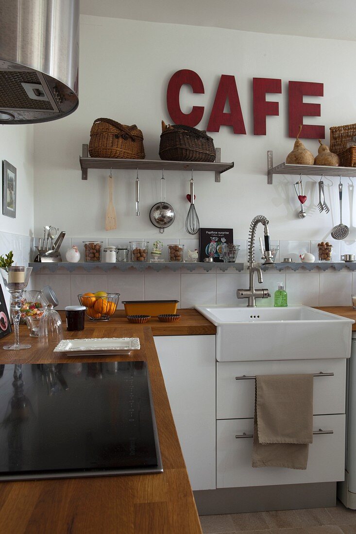 Holzarbeitsplatte und weiße Unterschränke, Wandkonsolen mit Küchenutensilien und dunkelroter Schriftzug in Küche mit Landhausflair