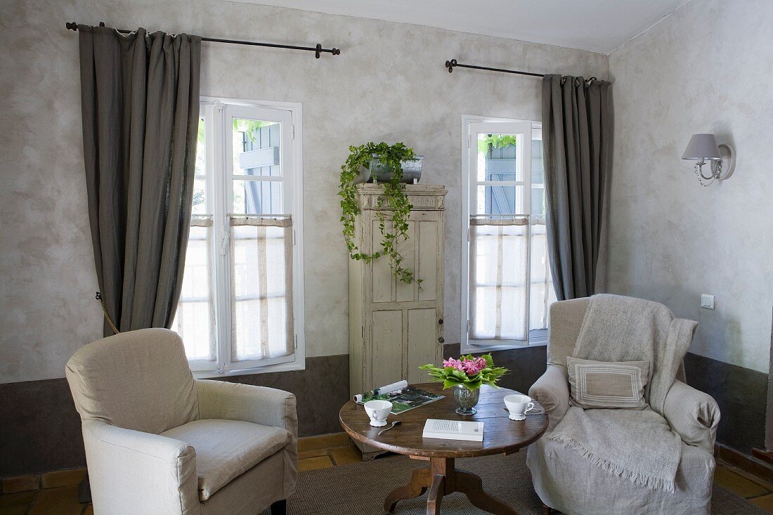 Zwei Sessel mit Hussen an rundem Biedermeiertisch und von Fenstern mit Vorhang flankierter, alter Schrank mit Efeupflanze