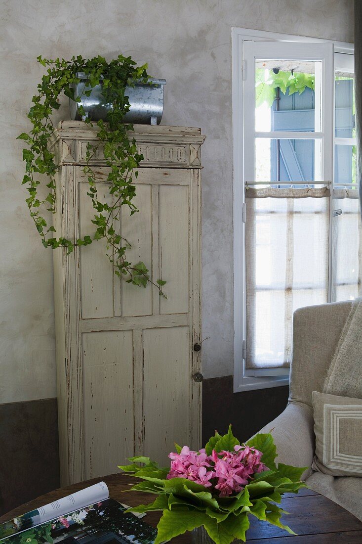 Zimmerefeu in Zinkgefäss auf antikem Schrank mit Patina, Bouquet mit Blättern und Blüten auf Tisch im Vordergrund