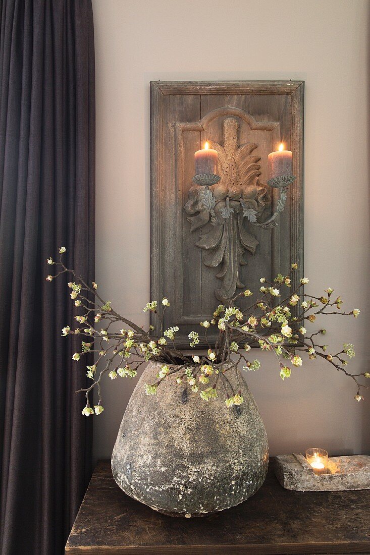 Vintage Vase mit Blütenzweigen auf Tisch, im Hintergrund Kerzenleuchter aus geschnitzter Holzplatte an Wand