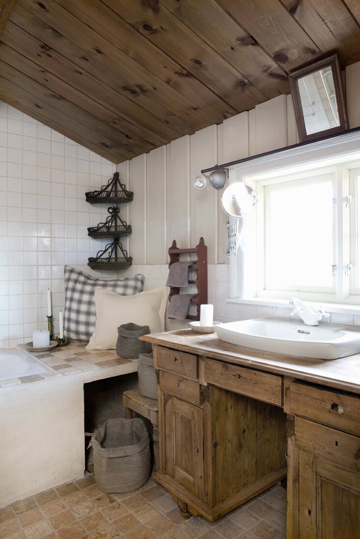 Rustikaler Waschtisch aus Holz vor Fenster, seitlich Kissen auf Ablage vor Badewanne
