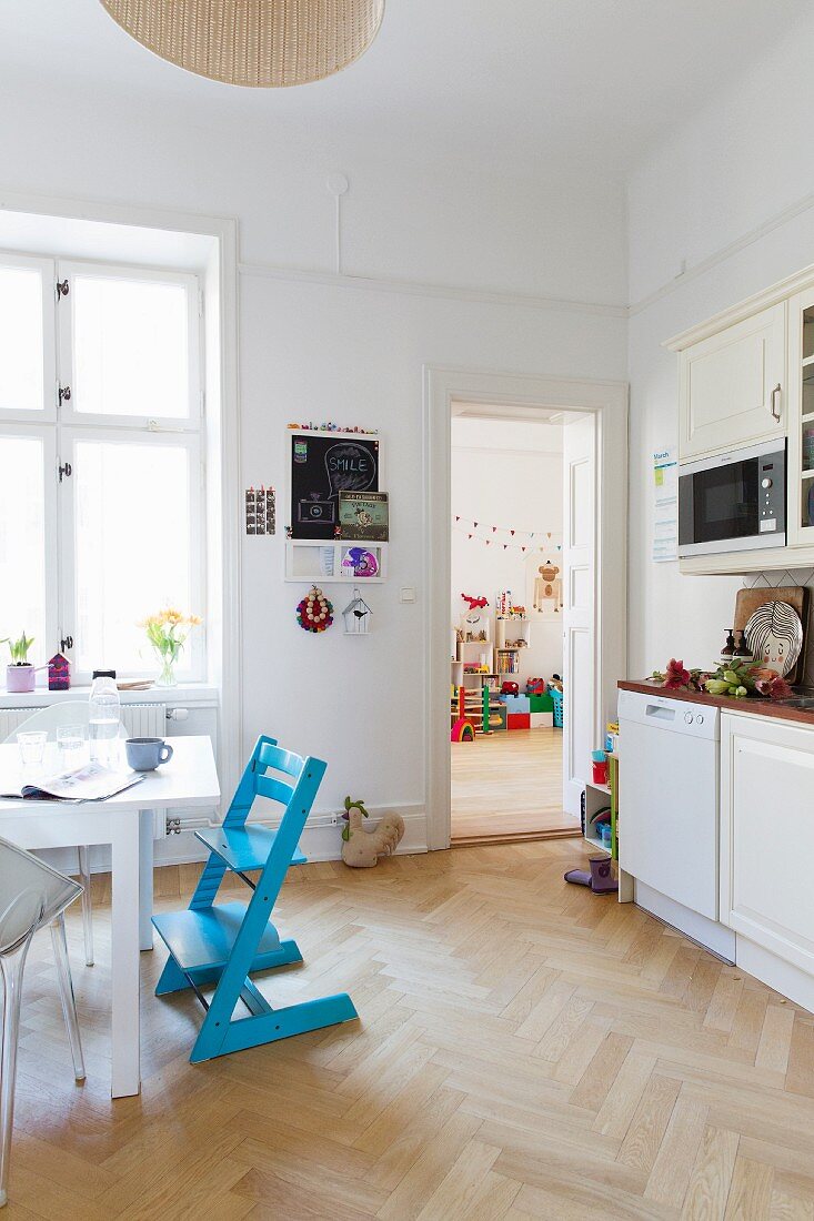 Esstisch und blauer Kinderstuhl in weisser Wohnküche; Blick in das Kinderzimmer im Hintergrund
