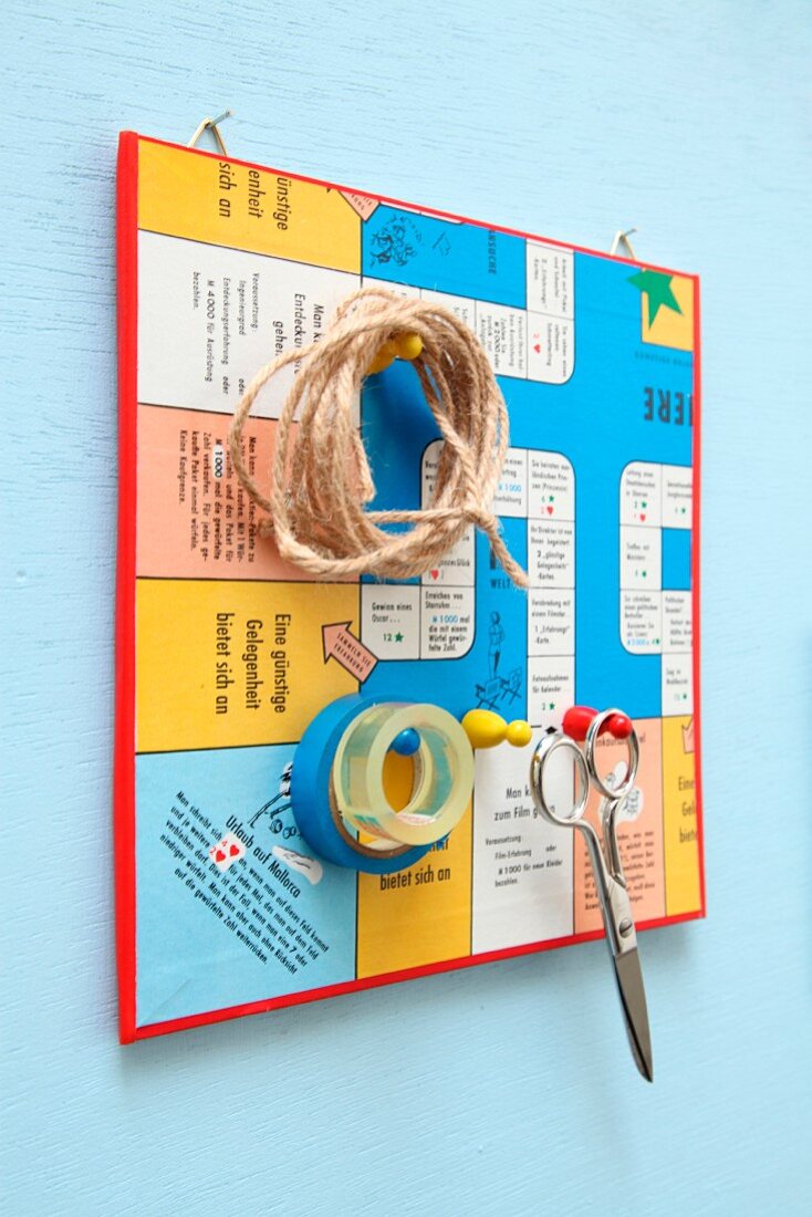Pinnwand aus einem zerschnittenen Brettspiel mit Spielfiguren als Aufhängungen für Schere, Schnur und Klebeband