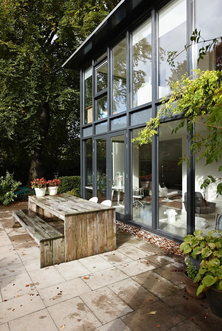 Zeitgenössisches Wohnhaus mit Glas-Stahlfassade, davor Terrassenplatz mit verwitterter Holztischgarnitur auf Fliesenboden