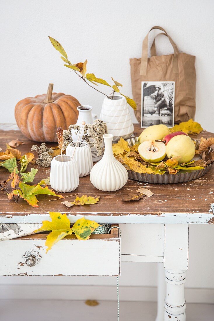 Vasensammlung aus weisser Keramik, zwischen Herbstlaub, Obst und Kürbis, auf ländlichem Holztisch