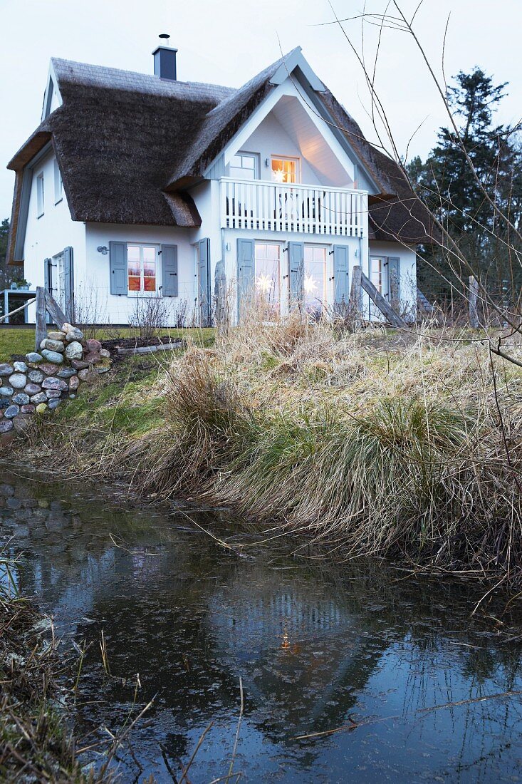 A small stream at the bottom of a garden with a thatched roof house in the background