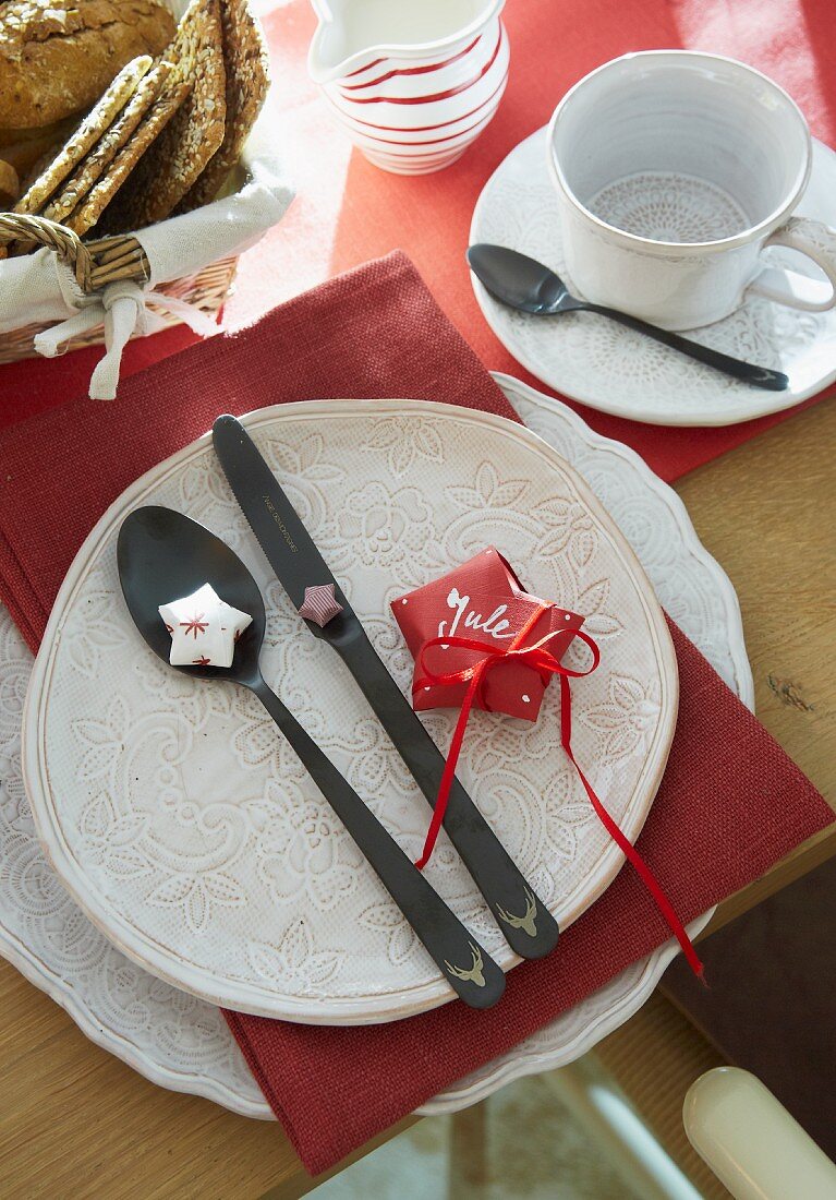 Roter Faltstern aus Papier als Platzkarte auf weißem Teller mit Besteck, daneben Tasse mit Untertasse