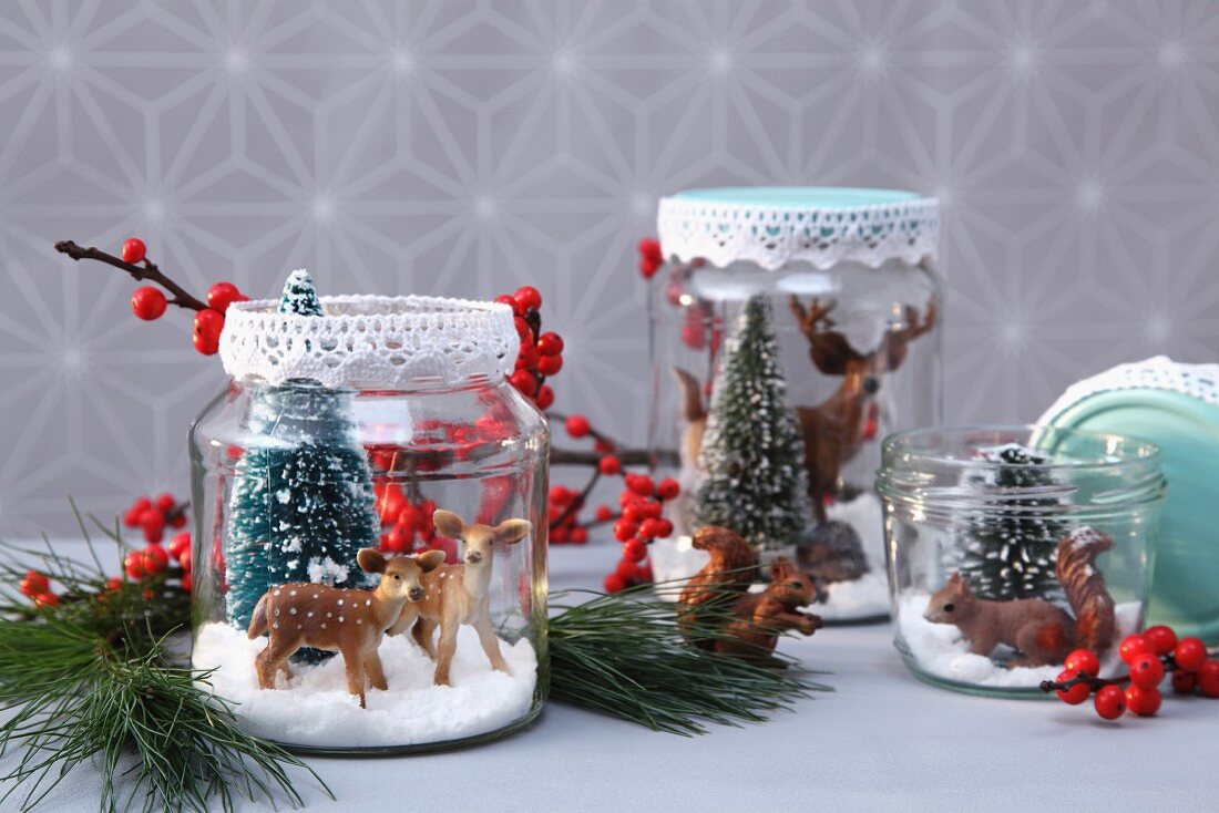 Plastiktiere und -tannen mit Kunstschnee in Konservengläsern, Deckel verziert mit Spitzenborte als weihnachtliche DIY-Deko