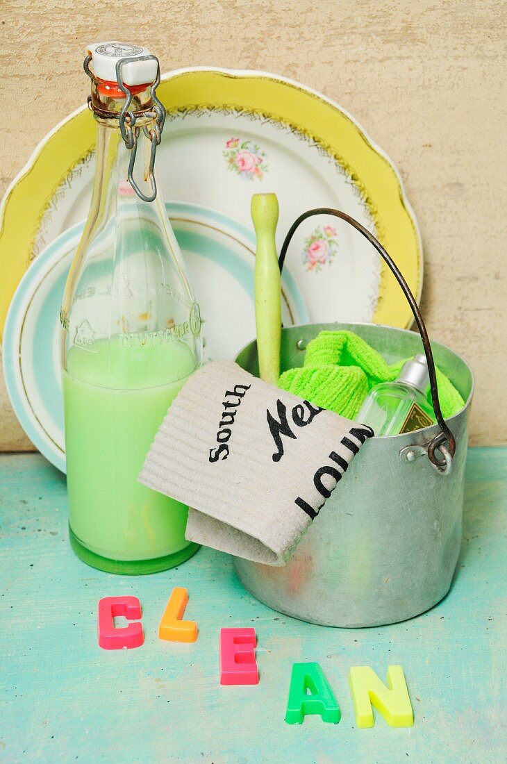 Vintage-style washing-up utensils with aluminium bucket, washing-up liquid in swing-top bottle, magnetic letters spelling 'Clean' and floral plates in background