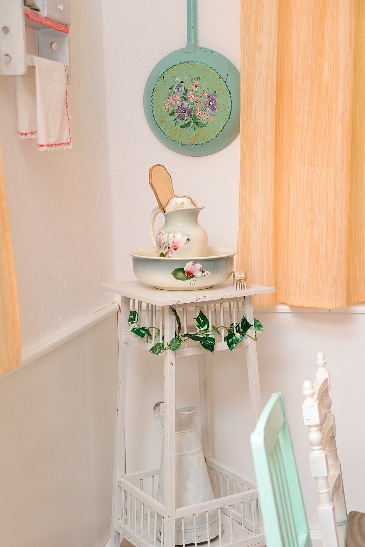 Ecke in Shabby Küche mit altem Waschset aus Porzellan und Emaille Kanne in Blumenständer, darüber eine bemalte Pfanne an der Wand