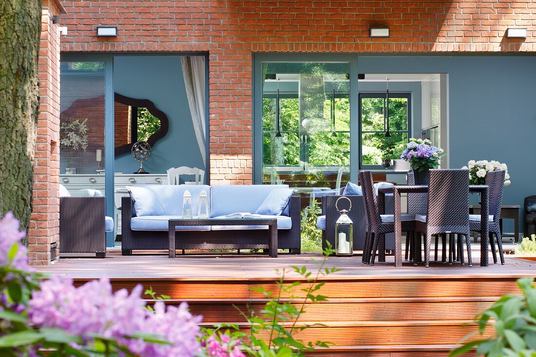 Dark, modern, wicker outdoor furniture on wooden deck in front of house with grey-painted window frames and wall elements in brick facade